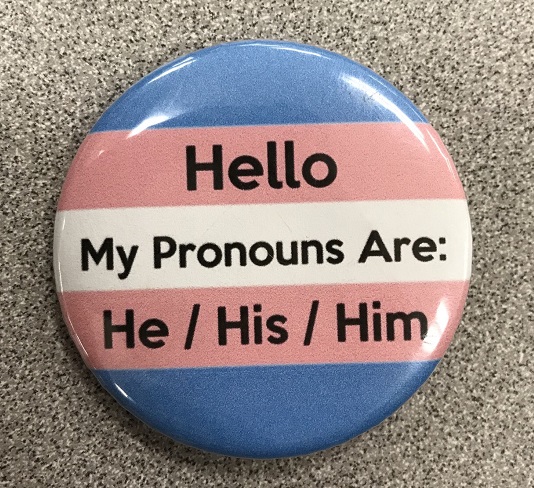 A pin that has the transgender flag on it as well as its wearers preferred pronouns