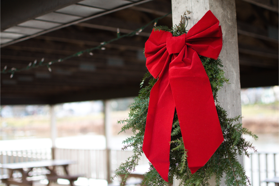 A wreath and bow placed on the wooden beams of a picnic area near the Susquehanna River downtown with string lights behind them, not yet illuminated.